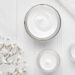 Open jars of white skincare cream on a clean white surface with small white flowers