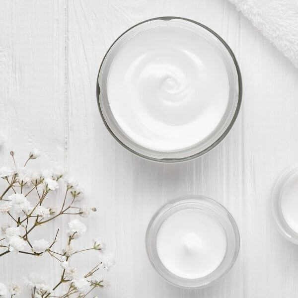 Open jars of white skincare cream on a clean white surface with small white flowers