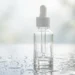 Clear glass serum bottle with white dropper on a dewy reflective surface